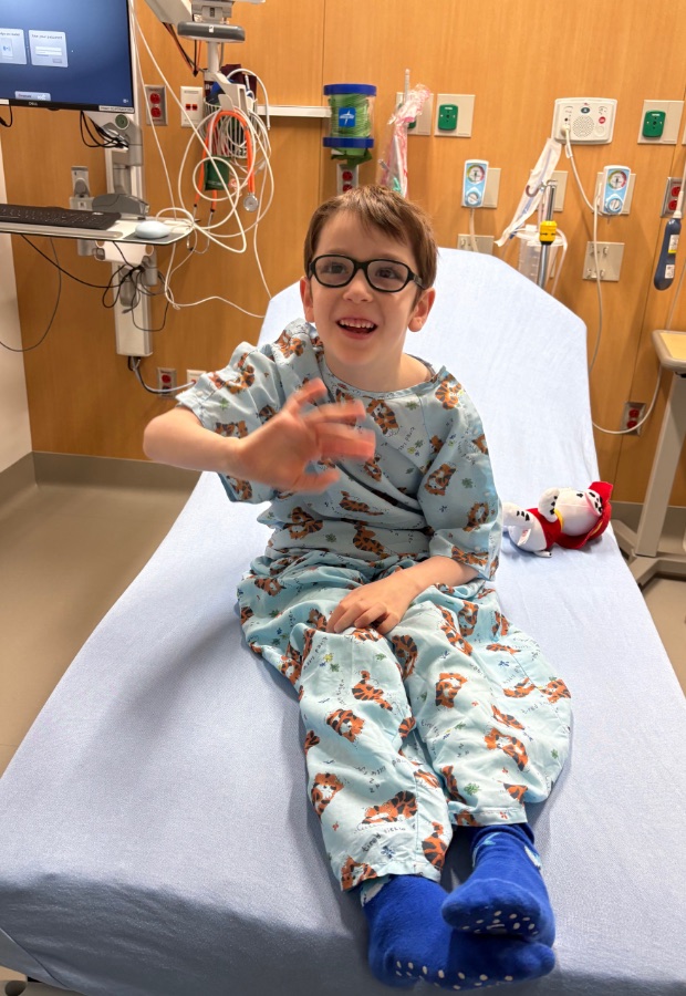 Alex in a hospital gown sitting on a bed, smiling and waving, before his brain MRI.