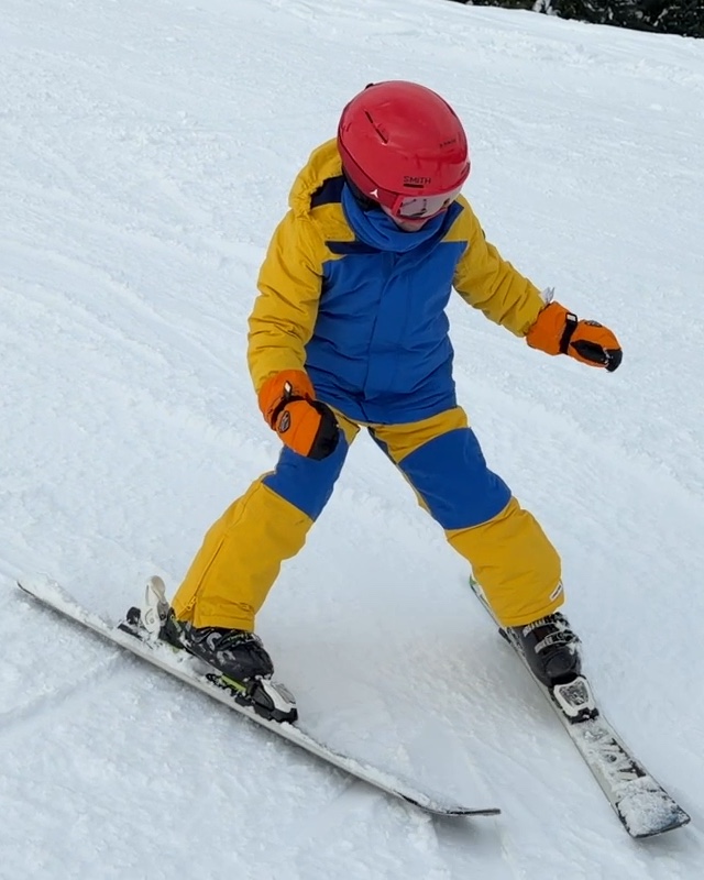 Alex skiing down a snowy slope in a red helmet, goggles and a yellow and blue snowsuit.