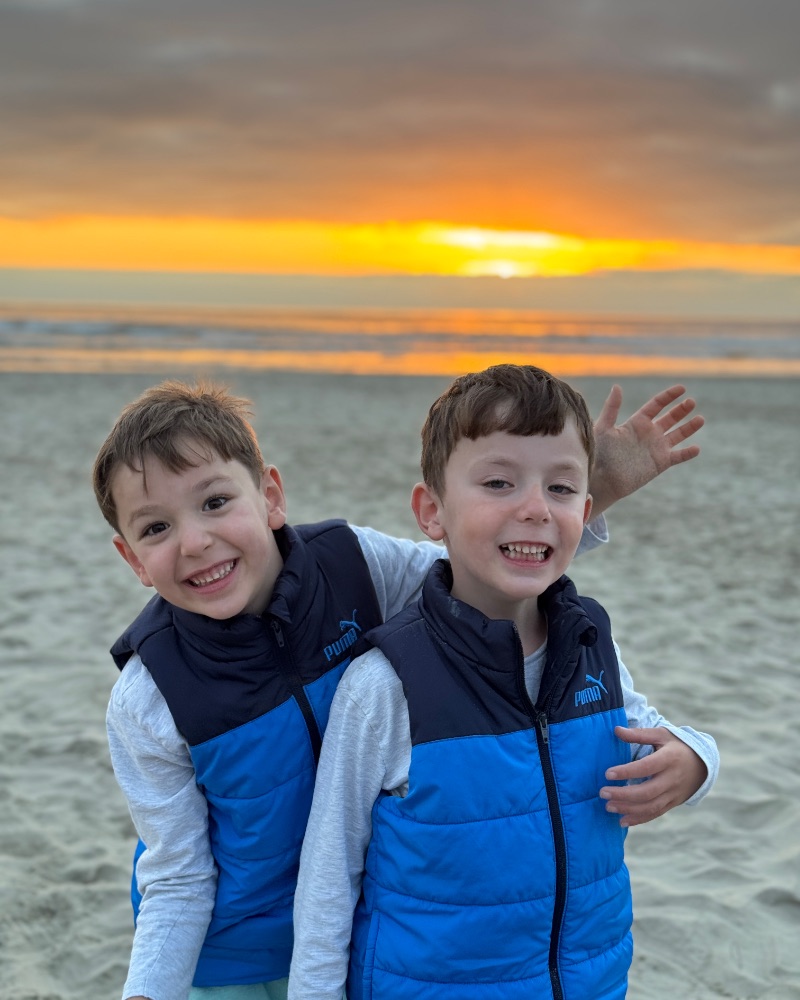 Alex and his twin brother Niko laughing together on a beach at sunset, Alex leaning in from the left.